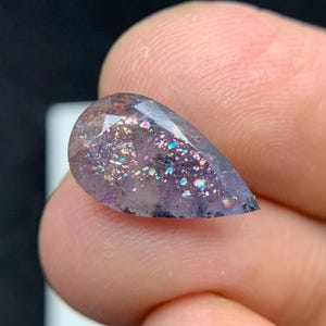 May include: A pear-shaped gemstone with a cut surface. The gem is a translucent purple colour with iridescent inclusions that reflect multiple colours. The gem is held between two fingers against a dark background.