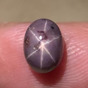 Puede incluir: Gema ovalada de color gris violáceo con un patrón de estrella de seis rayos. La superficie pulida presenta pequeñas imperfecciones. El efecto de estrella se crea por la reflexión de la luz en las inclusiones. Una pieza única y llamativa.