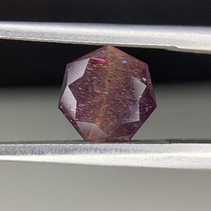 Op de afbeelding: Een gefacetteerde edelsteen met een complexe, geometrische vorm, vastgehouden door een pincet. De edelsteen vertoont een reeks kleuren, waaronder paars, roze en oranje, met interne insluitsels.