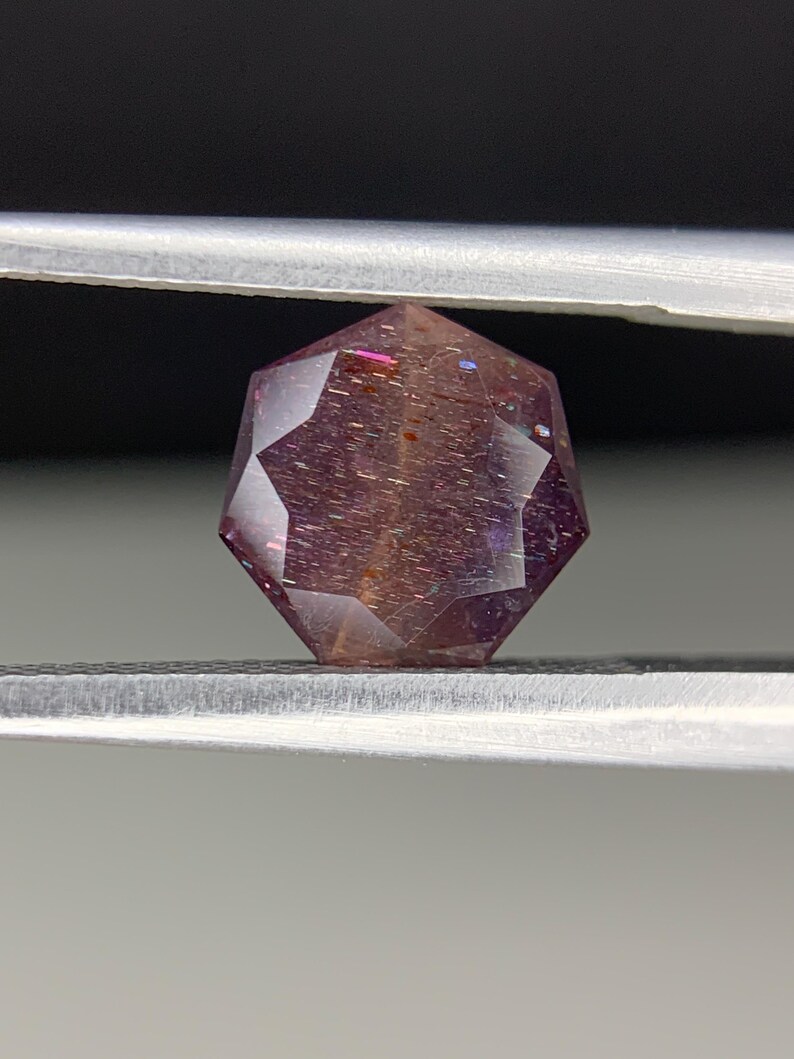 Op de afbeelding: Een gefacetteerde edelsteen met een complexe, geometrische vorm, vastgehouden door een pincet. De edelsteen vertoont een reeks kleuren, waaronder paars, roze en oranje, met interne insluitsels.