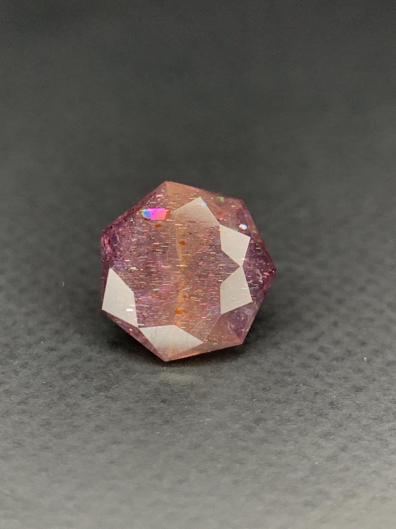 May include: A faceted, gemstone-like object with a complex geometric shape. The object has a reddish-purple hue with iridescent flashes of color. The surface reflects light, creating a sparkling effect. The gemstone is set against a dark background.