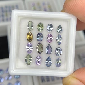 May include: A white square display box containing sixteen loose faceted gemstones arranged in a four-by-four grid. The stones feature various shapes including oval, pear, and marquise cuts, displaying a spectrum of colors such as green, yellow, purple, and blue.