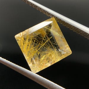 Pode incluir: Uma gema amarelo-dourada de corte quadrado com inclusões internas em forma de agulha, segurada por pinças prateadas contra um fundo escuro. As facetas da gema refletem a luz, mostrando sua clareza e padrões intrincados. Provavelmente é quartzo.
