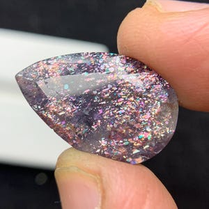 May include: A teardrop-shaped gemstone with a dark purple base and sparkling inclusions of various colours. The gem is held between fingers, showcasing its polished surface and iridescent effect. The gem's shape is smooth and rounded.