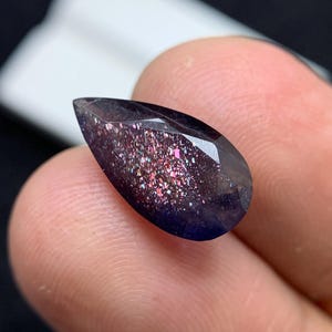 May include: A faceted, teardrop-shaped gemstone with a dark purple base and sparkling inclusions of pink, purple, and white. The gem is held between fingers, showcasing its polished surface and unique colour variations.