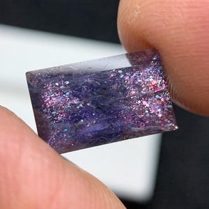 May include: A small, rectangular gemstone with a deep purple hue, speckled with iridescent glitter. The stone is held between two fingers, showcasing its polished surface and the play of light across its facets. The gem's edges are clean and well-defined.