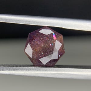 Op de afbeelding: Een gefacetteerde, veelkleurige edelsteen met een dieppaarse tint. De edelsteen wordt vastgehouden door een zilveren pincet tegen een donkere achtergrond. De facetten van de edelsteen reflecteren licht en cre&euml;ren een regenboogeffect.