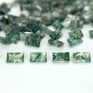 May include: A selection of rectangular moss agate gemstones. The stones are translucent with green, tree-like inclusions. The gemstones are scattered on a white surface, with a shallow depth of field.