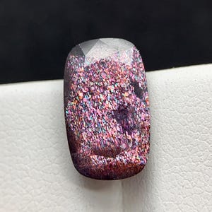 May include: A rectangular gemstone with a faceted top and a glittery, colourful interior. The gem's colours include shades of pink, purple, and blue, with a dark brown base. The gem is set against a white surface and a black background.