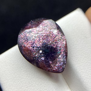 May include: A pear-shaped, faceted gemstone with a deep purple and dark red base colour. The surface displays a dense, shimmering aventurescence effect with scattered metallic-looking flecks that reflect light in shades of pink, blue, and silver.