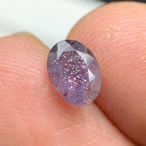 May include: An oval-cut gemstone with a speckled, shimmering appearance. The gem has a purple base colour with flecks of pink and silver, creating a glittery effect. The gem is held between two fingers.