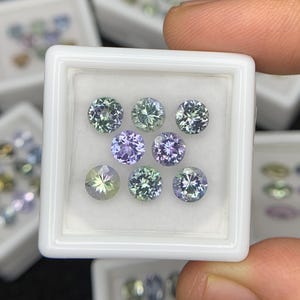 以下が含まれることがあります： 白いディスプレイケースに入った丸い宝石のコレクション。宝石は緑、青、紫など様々な色合いです。宝石はカットされており、光を反射しています。同様の宝石が入った他のディスプレイケースが背景にあります。
