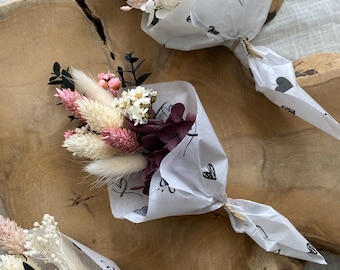 Puede incluir: Tres pequeños ramos de flores secas envueltos en papel blanco con diseños de corazones y garabatos negros. Los ramos presentan flores rosas, blancas y burdeos y vegetación. Los ramos están atados con hilo y descansan sobre una superficie de madera.