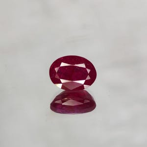 Op de afbeelding: Een gefacetteerde, ovaalvormige robijn edelsteen. De robijn heeft een dieprode kleur met interne facetten die licht reflecteren. De edelsteen wordt gepresenteerd op een reflecterend oppervlak, waardoor een spiegelbeeld ontstaat.