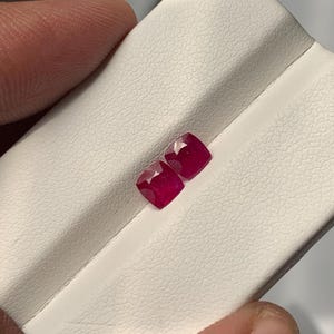 May include: Two cushion-cut, deep red rubies displayed on a white surface. The gemstones have multiple facets, reflecting light. The rubies are a vibrant, saturated color, showcasing their clarity and brilliance. The image highlights the gemstones' shape and color.
