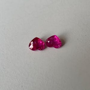 May include: Two heart-shaped, faceted gemstones in a vibrant magenta hue. The gems are displayed on a light grey surface, showcasing their polished surfaces and intricate cuts. These gems could be used for jewellery.