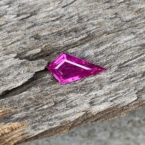 May include: A bright pink, faceted gemstone with a triangular shape. The gemstone is sitting on a piece of weathered wood.