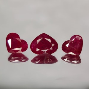 May include: Three heart-shaped, faceted ruby gemstones. The rubies are a deep red colour and have a high degree of clarity. The gemstones are displayed on a reflective surface, showcasing their brilliance and cut. The gems are likely for jewellery.