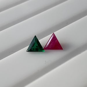 May include: Two triangular gemstones. One is a deep emerald green, the other a vibrant ruby red. Both are faceted and have sharp, clean edges. The gems are set against a white, ridged surface, creating a striking contrast.