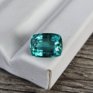 Puede incluir: Una gema verde vibrante con un corte de cojín, exhibida sobre una superficie blanca.