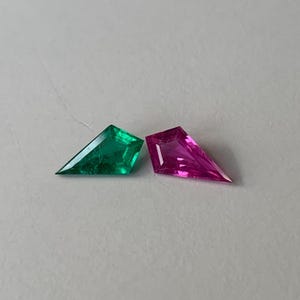 Puede incluir: Dos gemas con un corte geométrico único. Una es de un verde esmeralda vibrante, y la otra de un magenta profundo. Las gemas se muestran sobre una superficie gris claro, mostrando sus facetas. Ideal para la fabricación de joyas.