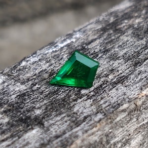 Op de afbeelding: Een levendige groene smaragd met een facetgeslepen vorm, liggend op een ruw, grijs houten oppervlak.