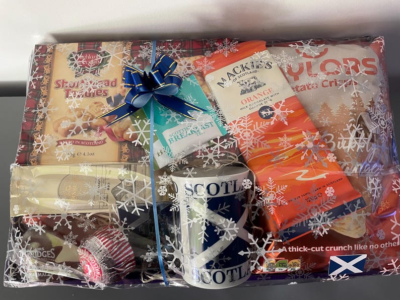 May include: A gift basket filled with Scottish themed items, including shortbread, tea, chocolate, crisps, and a mug with a Scottish flag design. The basket is wrapped in clear cellophane with a blue ribbon and snowflake pattern.