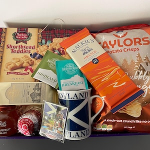 May include: A gift basket filled with Scottish themed items, including shortbread cookies, tea, chocolate, potato chips, and a mug. The basket is lined with red and green plaid fabric and has a blue and white Scottish flag mug.