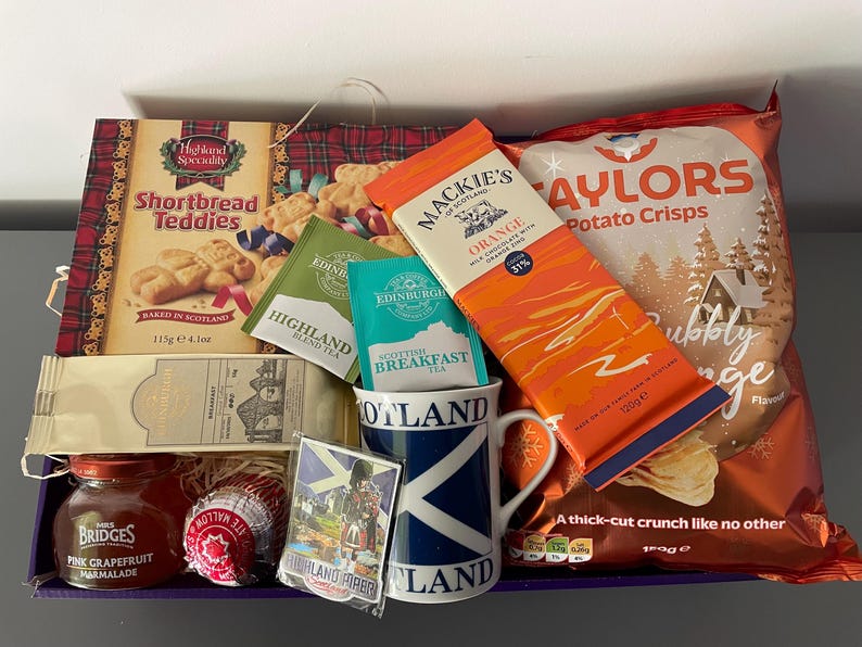 May include: A gift basket filled with Scottish themed items, including shortbread cookies, tea, chocolate, potato chips, and a mug. The basket is lined with red and green plaid fabric and has a blue and white Scottish flag mug.