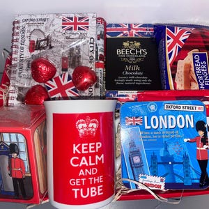 May include: A red and white gift basket with a mug that says "Keep Calm and Get the Tube" and a variety of British themed items, including chocolates, shortbread, and a London souvenir.