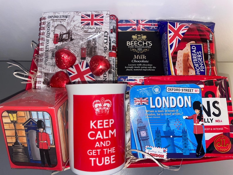 May include: A red and white gift basket with a mug that says "Keep Calm and Get the Tube" and a variety of British themed items, including chocolates, shortbread, and a London souvenir.