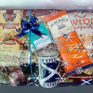 May include: A gift basket filled with Scottish themed treats, including shortbread, chocolate, and a mug with the Scottish flag. The basket is wrapped in clear cellophane with a blue ribbon and snowflakes.