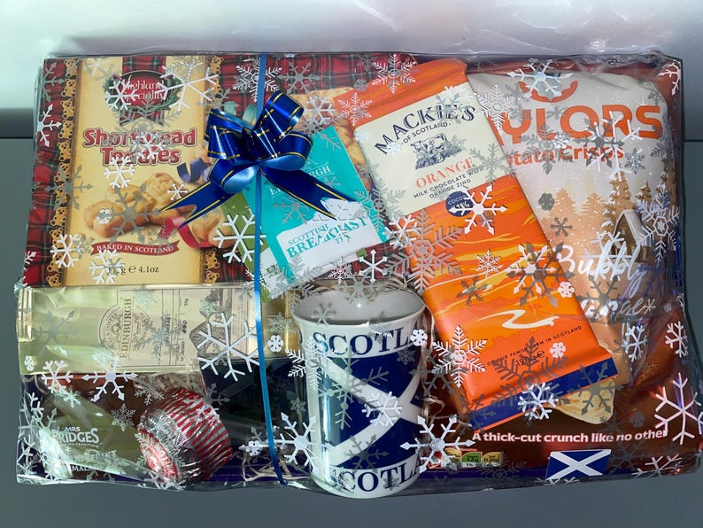 May include: A gift basket filled with Scottish themed treats, including shortbread, chocolate, and a mug with the Scottish flag. The basket is wrapped in clear cellophane with a blue ribbon and snowflakes.