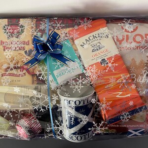 May include: A gift basket with a blue and white mug, a box of shortbread cookies, a box of orange chocolate, a box of biscuits, and a blue bow. The basket is wrapped in clear cellophane with a snowflake pattern.