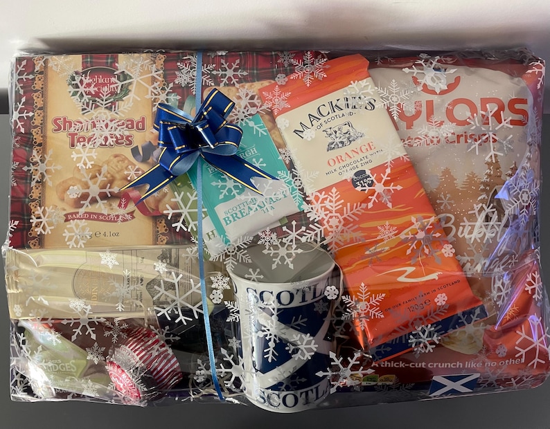 May include: A gift basket with a blue and white mug, a box of shortbread cookies, a box of orange chocolate, a box of biscuits, and a blue bow. The basket is wrapped in clear cellophane with a snowflake pattern.