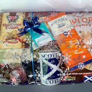 May include: A gift basket filled with Scottish themed treats, including shortbread cookies, orange milk chocolate, potato crisps, tea, and a mug with a Scottish flag design. The basket is wrapped in clear cellophane with a blue ribbon and snowflake decorations.