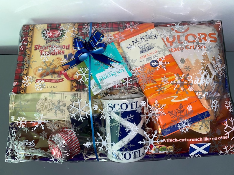 May include: A gift basket filled with Scottish themed treats, including shortbread cookies, orange milk chocolate, potato crisps, tea, and a mug with a Scottish flag design. The basket is wrapped in clear cellophane with a blue ribbon and snowflake decorations.