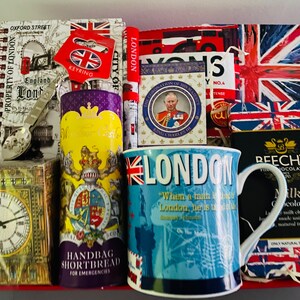 May include: A red and white gift basket filled with London themed items, including a mug, a notebook, a box of shortbread, and a box of chocolates. The mug has a blue background with the word "London" in white letters and a quote about the city. The notebook has a map of London on the cover. The shortbread box has a picture of Big Ben on the front. The chocolate box has a picture of the Union Jack flag on the front.
