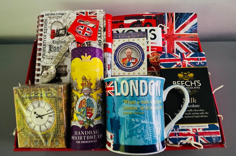 May include: A red and white gift basket filled with London themed items, including a mug, a notebook, a box of shortbread, and a box of chocolates. The mug has a blue background with the word "London" in white letters and a quote about the city. The notebook has a map of London on the cover. The shortbread box has a picture of Big Ben on the front. The chocolate box has a picture of the Union Jack flag on the front.