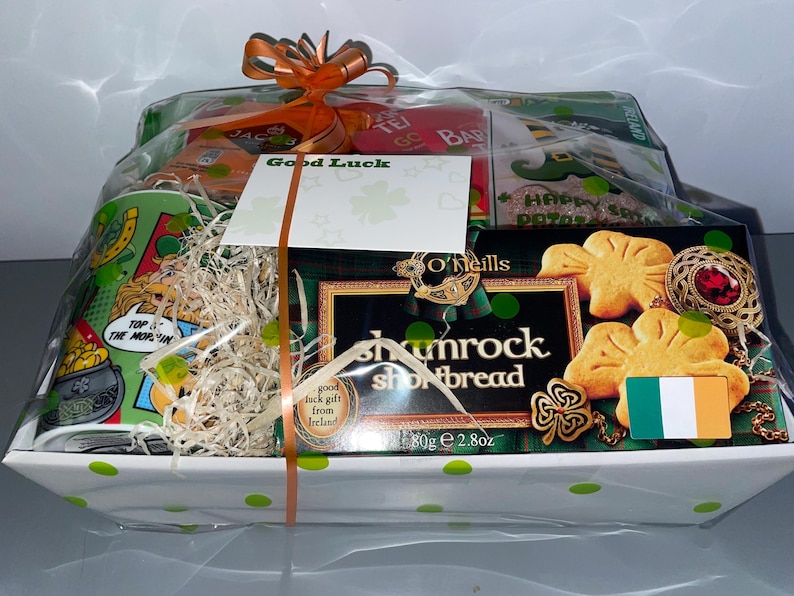 May include: A white gift basket with green polka dots filled with Irish themed gifts. The basket includes a mug with a cartoon image of a leprechaun, a box of O'Neill's Shamrock Shortbread, a bag of tea, and a card that says "Good Luck".