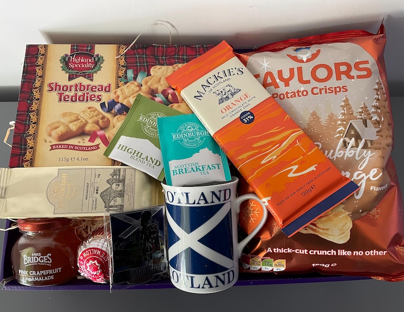 May include: A gift basket filled with Scottish themed items, including shortbread cookies, tea, chocolate, potato chips, and marmalade. The basket is lined with a red and black plaid fabric and features a blue and white mug with the word "Scotland" printed on it.