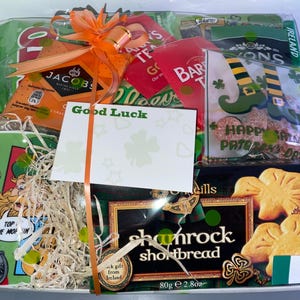 May include: A gift basket filled with Irish-themed snacks and treats. The basket includes a mug, tea bags, shortbread cookies, and a card that says "Good Luck".