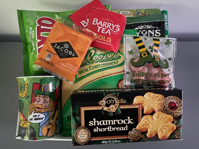 May include: A green and gold gift basket filled with Irish themed snacks and treats. The basket includes a mug with a cartoon leprechaun, a box of shamrock shortbread, a bag of cream crackers, a bag of candy, and tea bags.