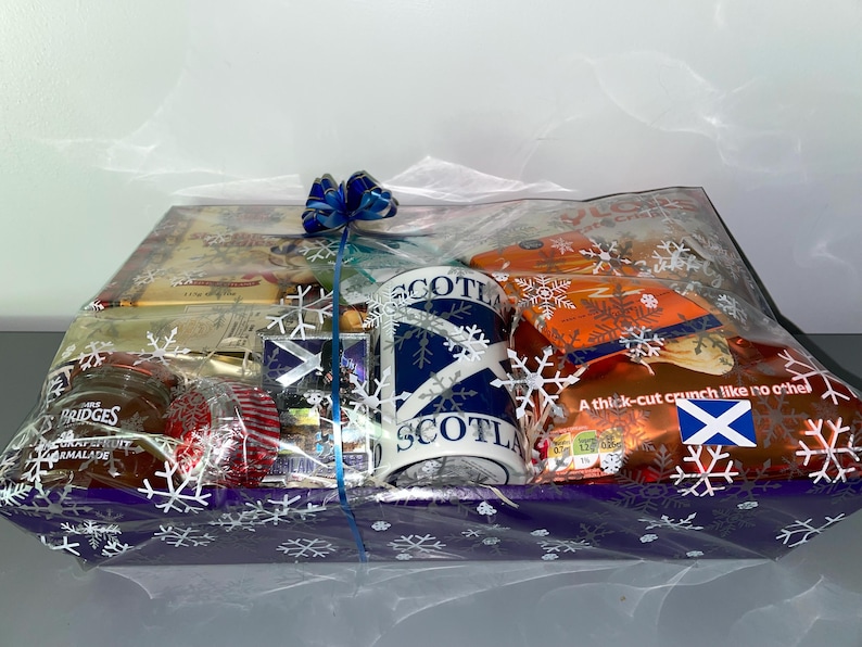 May include: A purple and white gift basket filled with various Scottish themed items, including a mug with the word "Scotland" on it, a bag of crisps, and a jar of marmalade. The basket is wrapped in clear cellophane and tied with a blue ribbon.