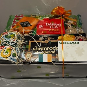 May include: A white gift basket filled with Irish themed items, including a mug with a shamrock design, tea, shortbread, and a card that says "Good Luck".