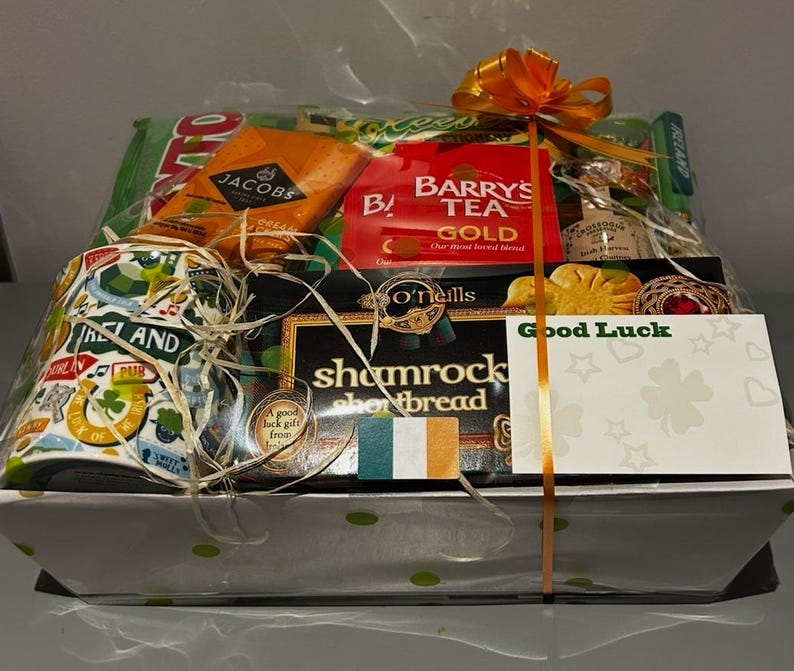 May include: A white gift basket filled with Irish themed items, including a mug with a shamrock design, tea, shortbread, and a card that says "Good Luck".