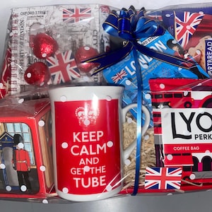 May include: A red and white gift basket with a mug that says "Keep Calm and Get the Tube" and a bag of Lyons Perkadilly coffee. The basket also includes a small box with a picture of a red telephone booth and a British guard, and a package of gingerbread fingers.