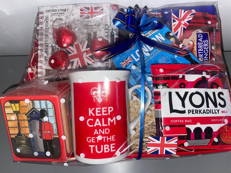 May include: A red and white gift basket with a mug that says "Keep Calm and Get the Tube" and a bag of Lyons Perkadilly coffee. The basket also includes a small box with a picture of a red telephone booth and a British guard, and a package of gingerbread fingers.