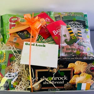 May include: A green and white gift basket filled with Irish themed items. The basket includes a mug, tea, shortbread cookies, candy, and a card that says "Good Luck".