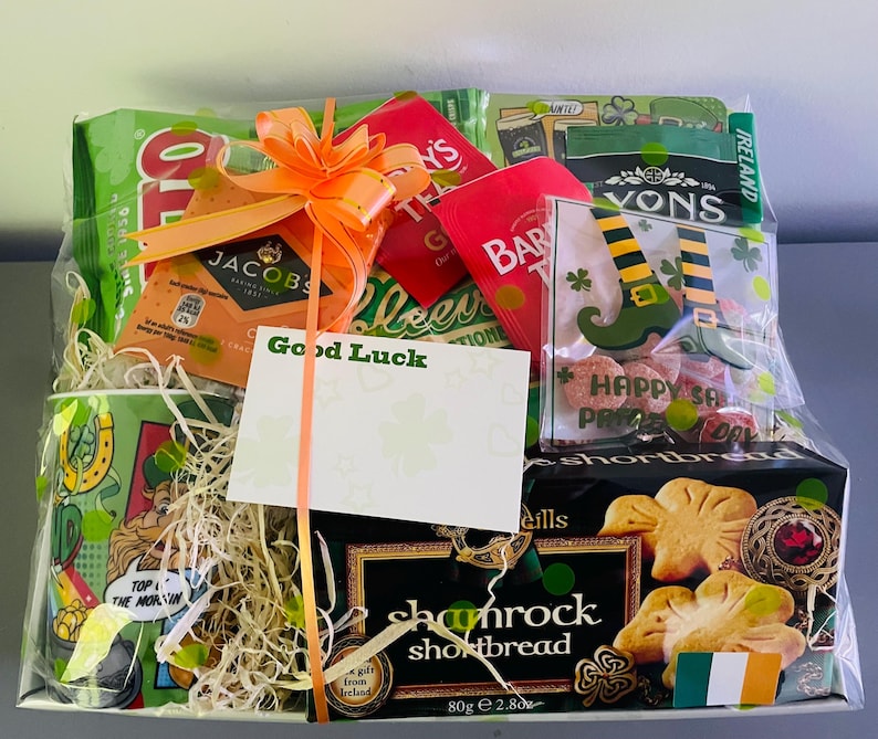 May include: A green and white gift basket filled with Irish themed items. The basket includes a mug, tea, shortbread cookies, candy, and a card that says "Good Luck".